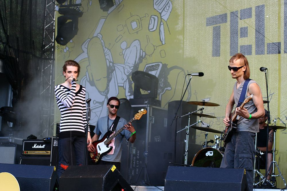 Four musicians perform on an outdoor stage with a yellow backdrop featuring abstract art at Positivus AB 2008. The lead singer, wearing a striped shirt, holds a microphone, while a guitarist in sunglasses and a sleeveless shirt stands to his right. A bassist and drummer are at the back, playing their instruments.