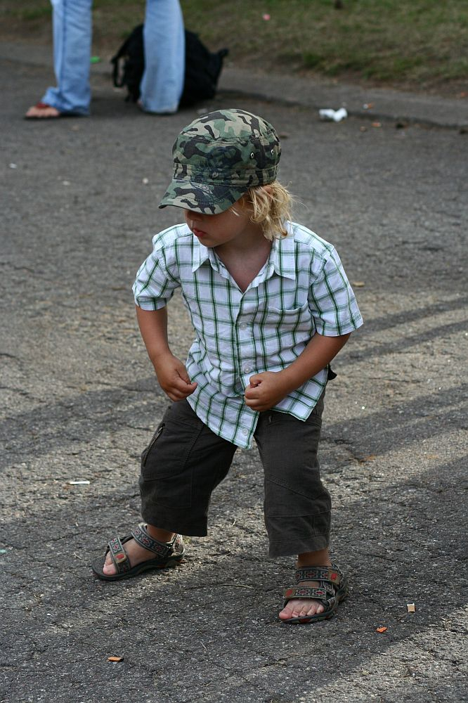 A young child with blonde hair stands on a paved surface, wearing a green camouflage cap, a white and green plaid short-sleeve shirt, dark pants, and open-toed sandals. The child is bending slightly forward with both hands on their knees. A black backpack from Positivus AB 2008 is in the background.