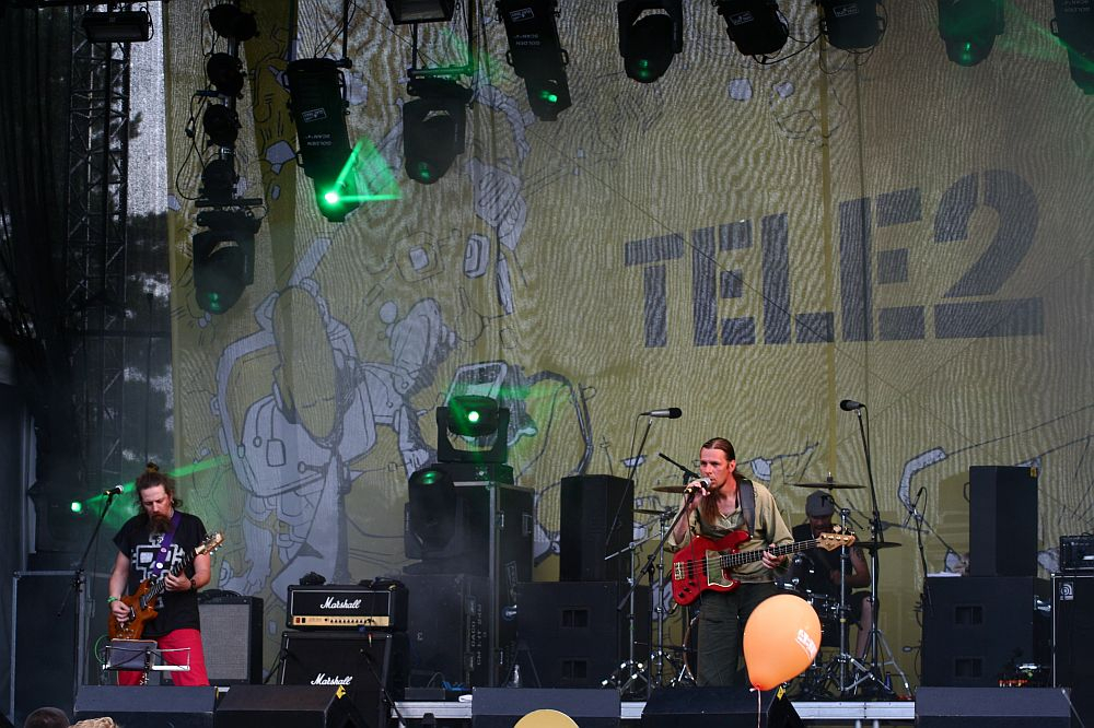 Three musicians perform on a stage with various instruments, including a guitar and bass, under the AB 2008 banner. The backdrop features the "Tele2" logo surrounded by abstract art. Multiple stage lights illuminate the performers while yellow balloons float near the stage, capturing a Positivus outdoor concert vibe.
