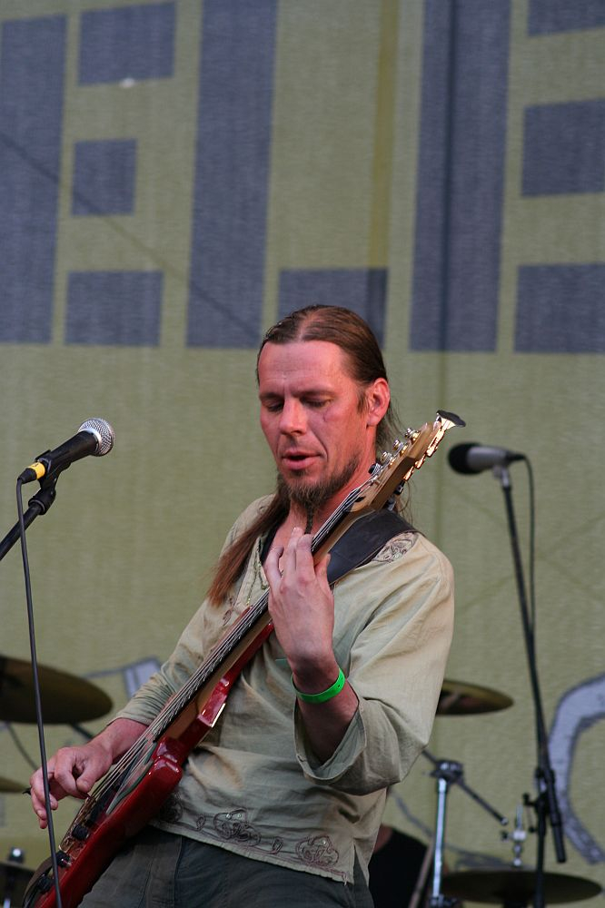 At Positivus 2008, a musician with long hair tied back plays a red bass guitar on stage. He wears a light green shirt and has a focused expression. A microphone is set up in front of him, and a drum set is visible in the background. The stage backdrop features large, partially visible letters, possibly "AB.