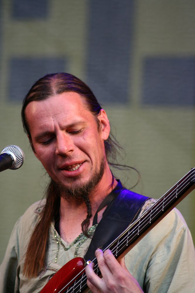 At AB 2008, a man with long hair tied back and a small beard and mustache plays an electric bass guitar on stage. Wearing a beige shirt, he appears focused on his performance, with a microphone in front of him. The background is blurred, indicating stage lighting and possibly a Positivus banner.