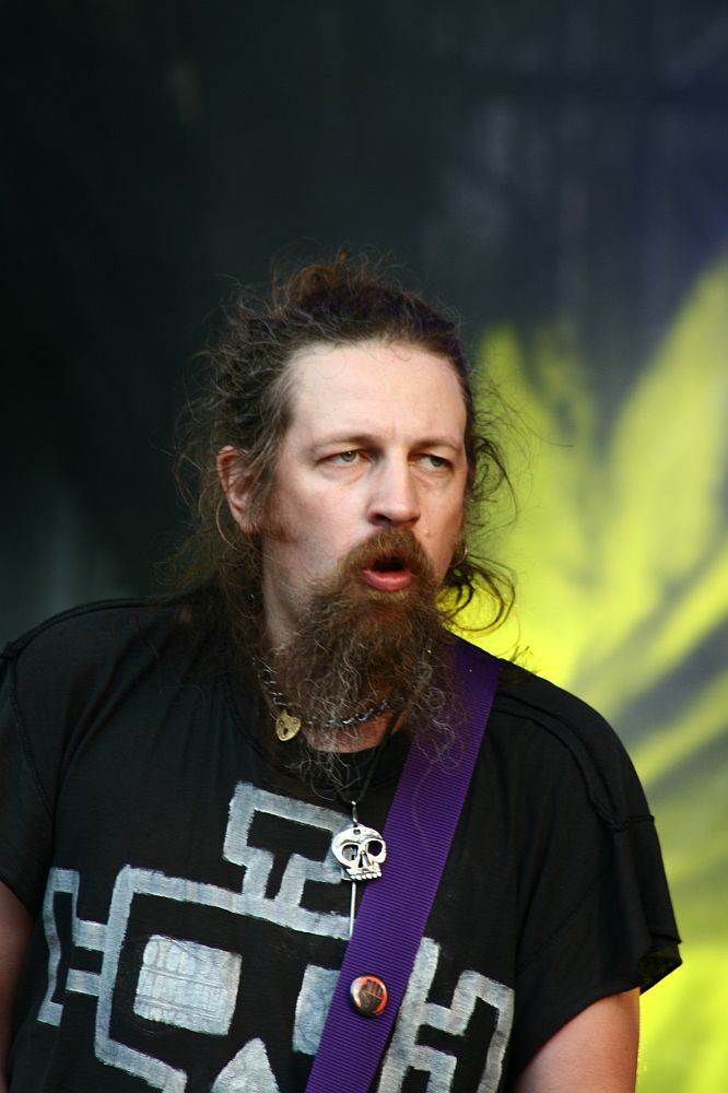At Positivus 2008, a man with long hair and a beard stands on stage, wearing a black shirt with a white geometric design and a purple guitar strap. He has multiple necklaces, including one with a skull pendant, and appears to be singing or speaking against the dramatic yellow lighting backdrop.