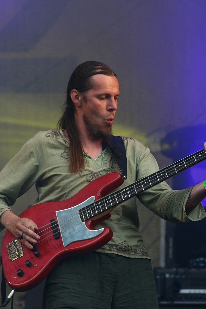 A musician with long hair, wearing a light green shirt, plays a red electric bass guitar. He appears to be deeply focused and immersed in the music, with stage lighting in hues of purple and yellow. The setting suggests a live performance at Positivus AB 2008 festival.