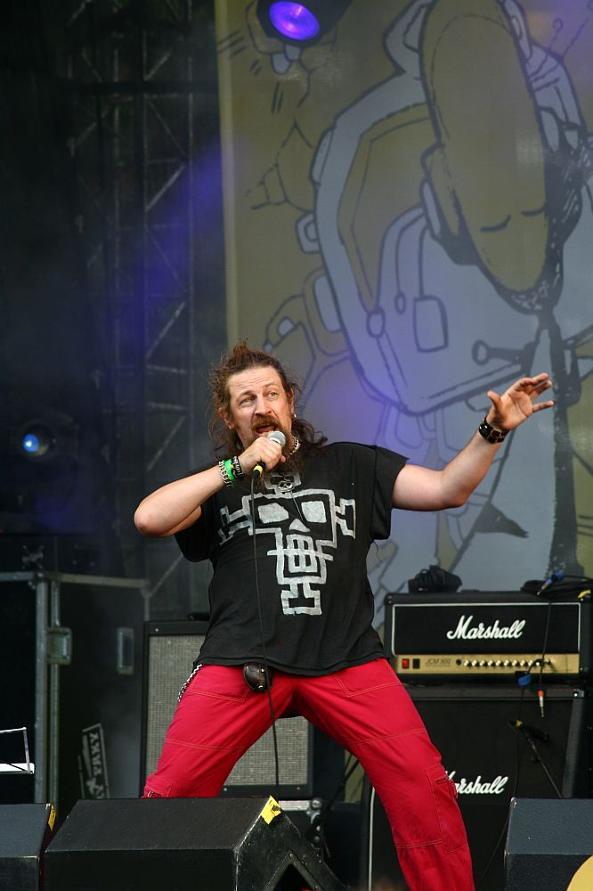 A person with long hair and a beard passionately sings into a microphone on stage at Positivus. They wear a black t-shirt with a white graphic and red pants. Behind them, a large banner featuring abstract art reads "AB 2008," while amplifiers and vibrant stage lighting add to the electric atmosphere.