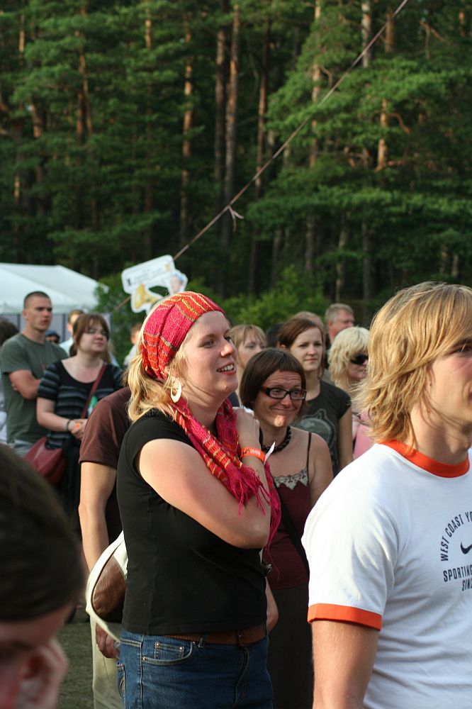 A diverse group of people stands outdoors at a forested location, attending an event reminiscent of Positivus. In the foreground, a woman wearing a colorful headscarf and earrings smiles while looking ahead. Other attendees, casually dressed, are visible in the background among trees and signs with AB 2008 references.