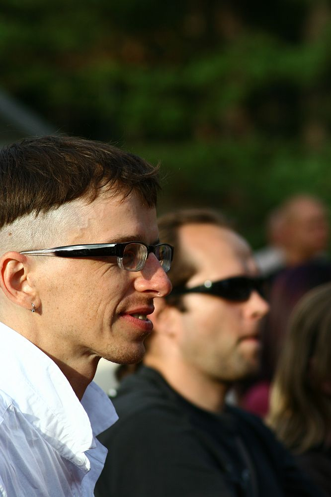 A person with short, dark hair and a shaved section wearing glasses and a white shirt is facing left in an outdoor setting at Positivus 2008. Another person with short dark hair and sunglasses is seen in the background, slightly out of focus. Trees or foliage are blurred in the background.