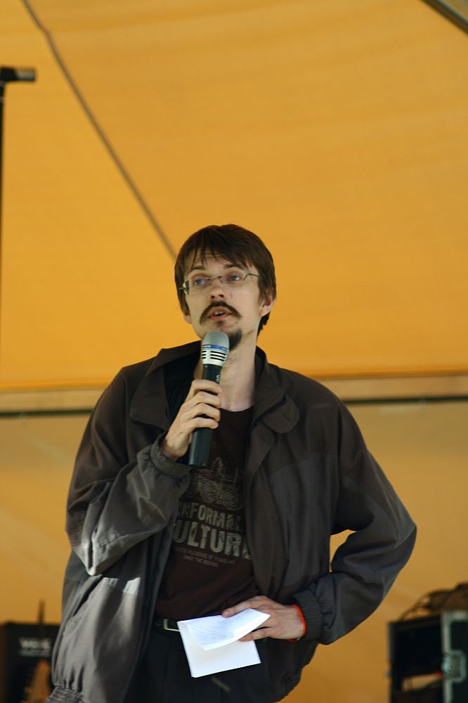 A man with a mustache and glasses is holding a microphone and a piece of paper, speaking under a yellow canopy at the AB 2008 event. He is wearing a dark jacket over a t-shirt with indistinguishable text. His other hand rests on his hip, exuding confidence while Positivus equipment is visible in the background.