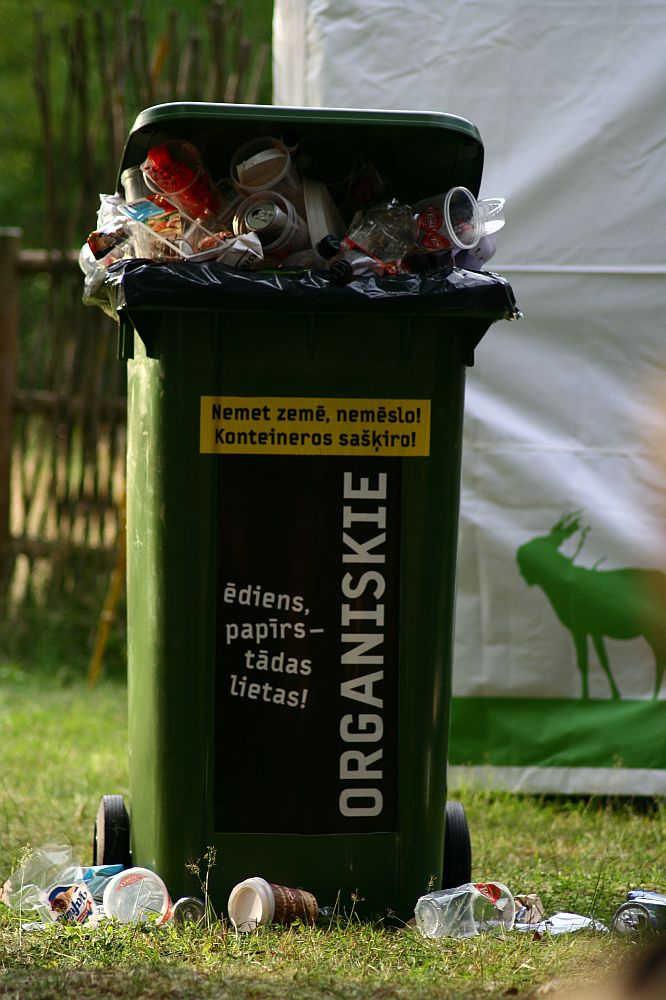 A green trash bin labeled "ORGANISKIE" at the 2008 Positivus festival is overflowing with various waste items such as plastic cups, cans, and paper. Additional litter surrounds the base of the bin. There is a white and green sign in the background and a wooden fence partially visible to the left. The text on the bin reads: "Nemē tī zemi, nemet