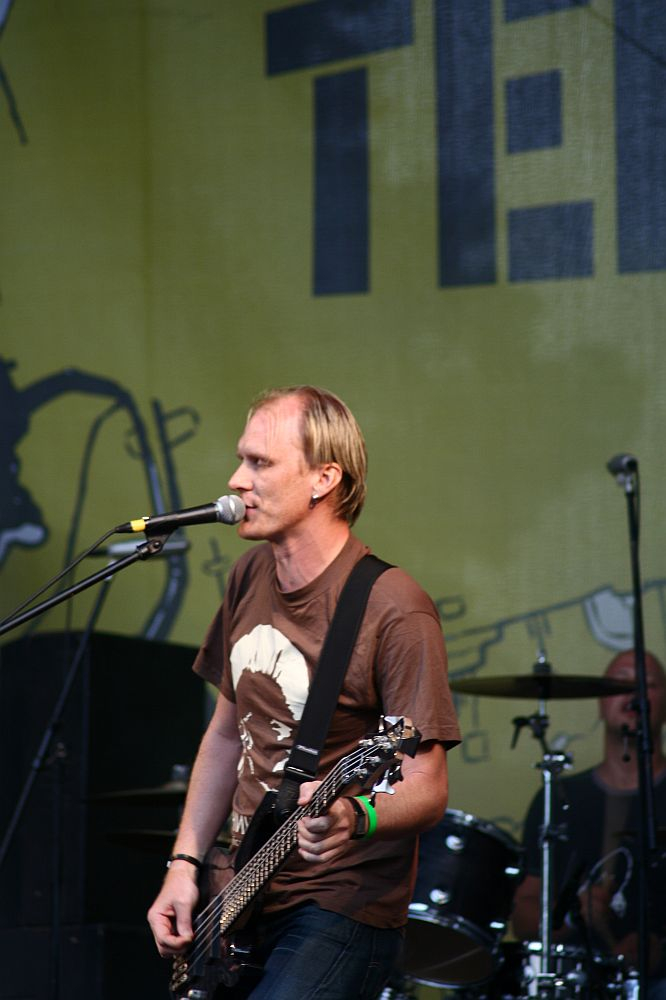 A man is on stage at Positivus, playing an electric guitar and singing into a microphone. He is wearing a brown T-shirt and blue jeans. Behind him, a drummer is partially visible, playing the drums. The background features a large, yellow banner with indistinct black graphics and text, reminiscent of AB 2008.