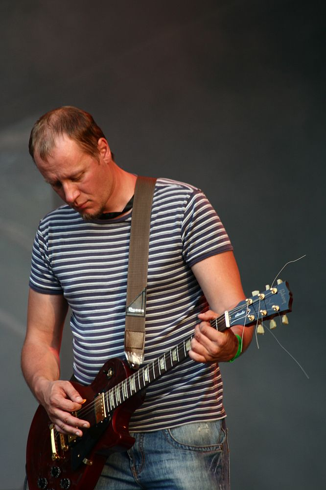 A man with short hair, wearing a striped t-shirt and jeans, is playing a brown electric guitar at the Positivus festival. He stands against a blurred dark background, looking down at the guitar, focused on his performance. His guitar strap is firmly placed on his shoulder.