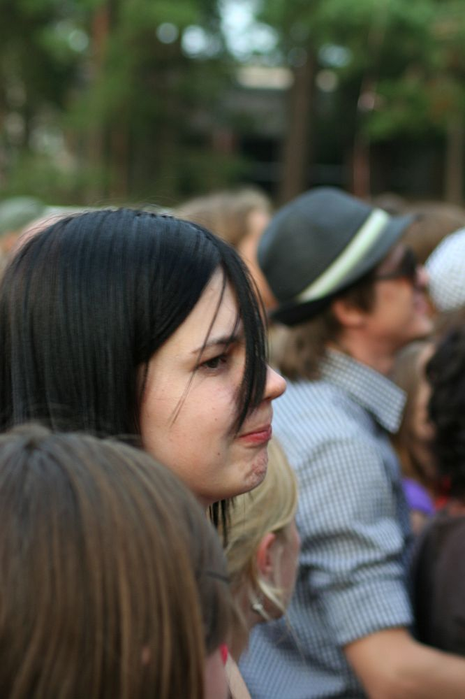 A person with long black hair looks to the side in a crowded outdoor setting at Positivus. They are surrounded by others, including a person in a gray fedora. The background shows blurred trees and people, indicating a possible event or gathering. The person appears thoughtful or pensive.