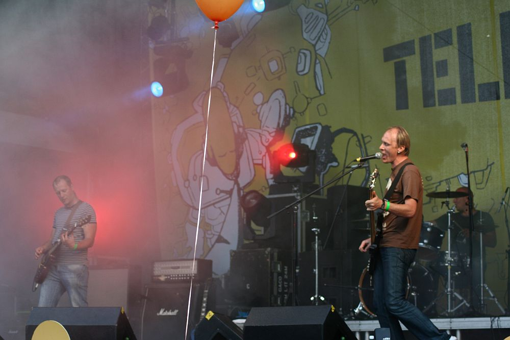 A band performs on an outdoor stage at Positivus. The bassist, wearing a striped shirt, and the vocalist, in a brown shirt, play guitar and sing into a microphone. A drummer is visible in the background. Bright lights, smoke effects, and a yellow backdrop with illustrations and text add to the dynamic scene.