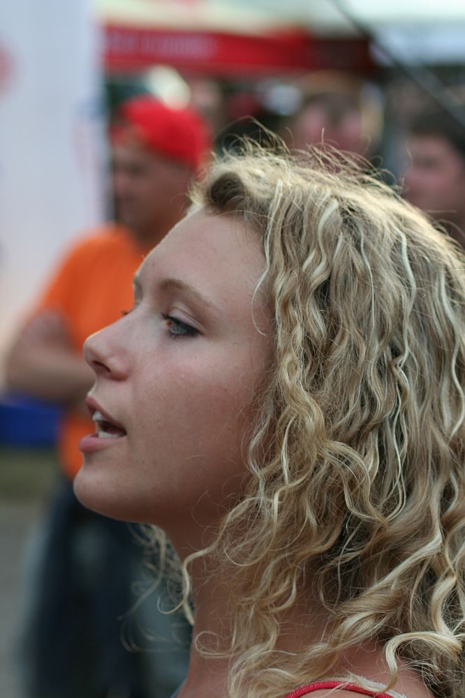 A young woman with long, curly blonde hair, likely at Positivus AB 2008, is shown in side profile, facing left. She appears to be singing or speaking. The background is blurry with people standing and possibly some outdoor event tents. Dominant colors in the background are red, orange, and white.