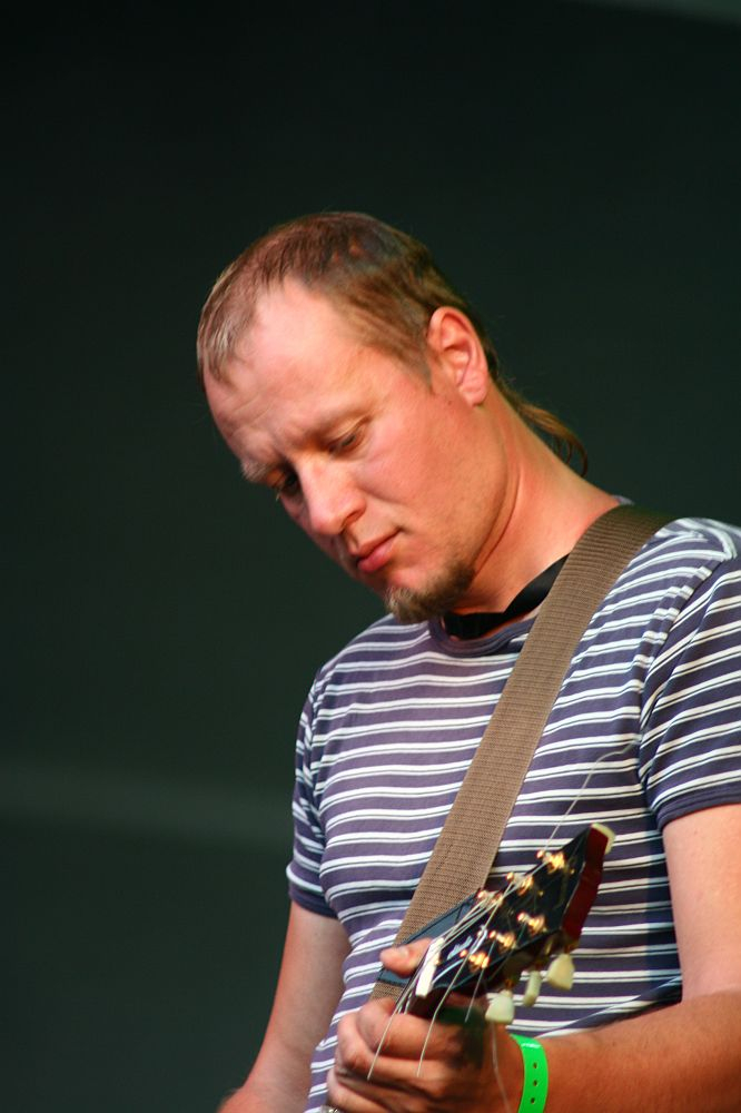 A person with short, light brown hair and a short beard is performing on stage at Positivus, concentrating on playing an electric guitar. They are wearing a striped short-sleeved shirt and a green wristband. The background is a plain, dark color, reminiscent of AB 2008 vibes.
