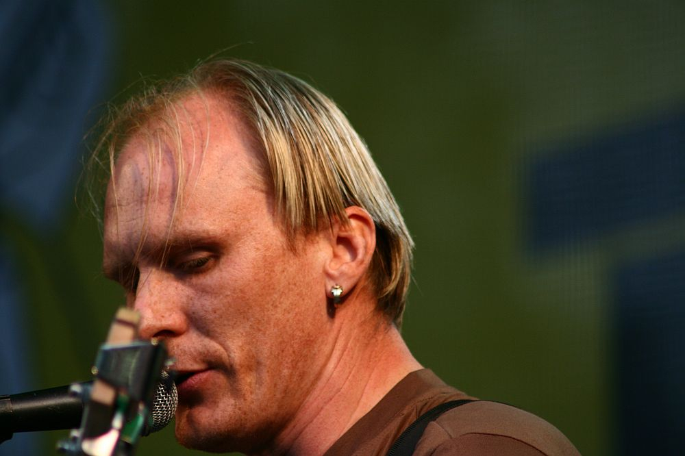 A person with short blonde hair and an earring is shown in close-up, singing into a microphone at Positivus AB. They are wearing a brown shirt and focused on performing, with a greenish-yellow blurred background suggesting an outdoor setting. The image captures a moment of intensity and concentration from 2008.