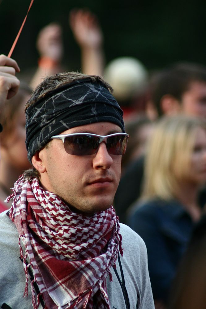 Amidst the vibrant crowd at Positivus, a person wearing dark sunglasses, a black headband, and a red and white patterned scarf stands out. Dressed in a white or light-colored shirt, they blend with the blurred figures in the background raising their arms, likely enjoying an event or concert reminiscent of 2008.