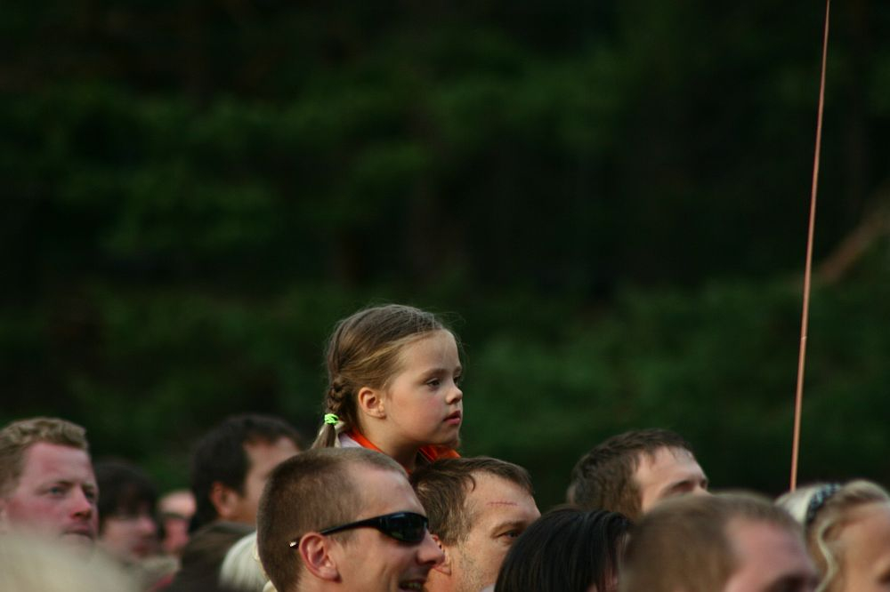 A young child with blond hair in braids sits on an adult's shoulders in a crowd at Positivus AB 2008. The child has a focused expression and is wearing an orange shirt. The surrounding adults have varied expressions and appearances, with green foliage from trees visible in the background.