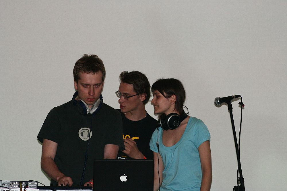 Three individuals are focused on an audio setup at a Positivus 2008 event. The person on the left is adjusting equipment while wearing headphones and looking serious. The person in the middle is speaking to the others. The woman on the right is smiling and also wearing headphones. A microphone stand is visible to the right.