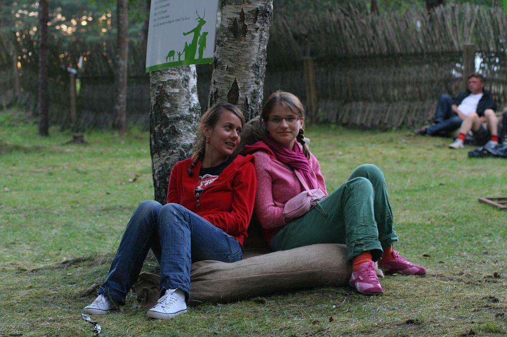 Two young women are sitting on a burlap sack in a grassy outdoor area at Positivus, leaning against a tree. Both are wearing casual clothes; one in a red hoodie, the other in a pink hoodie. They are looking at something off to the side. A few other people are visible in the background, seated near a wooden fence.