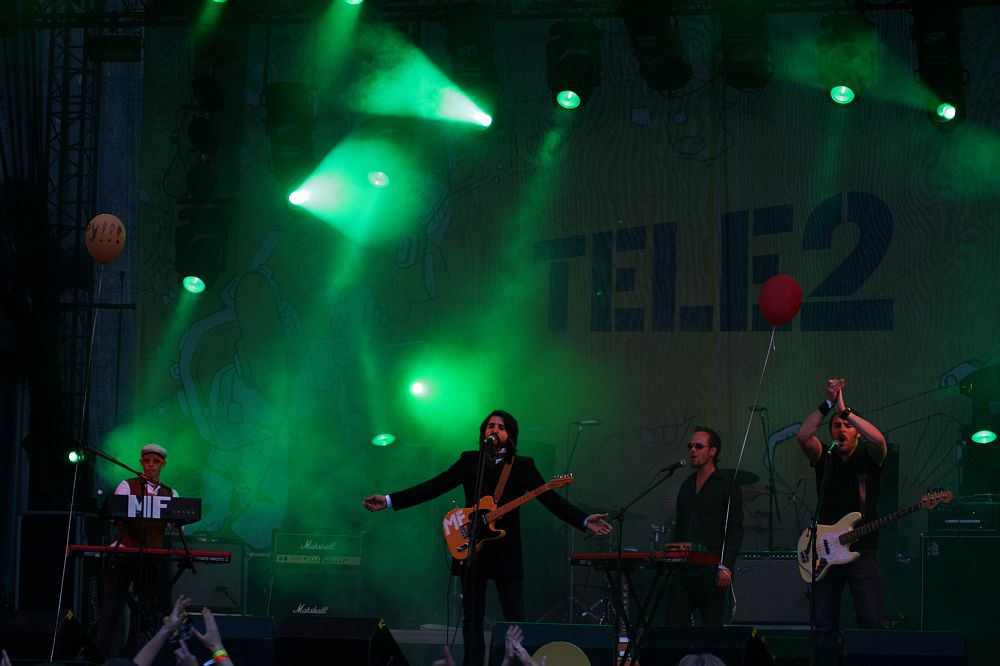 Band performing on a stage with green lighting at Positivus AB, 2008. The lead singer, in the center, holds a guitar and sings into a microphone. Two musicians on either side play keyboards, and one plays a bass guitar. The backdrop displays "TELE2." Balloons float above the partially visible crowd.