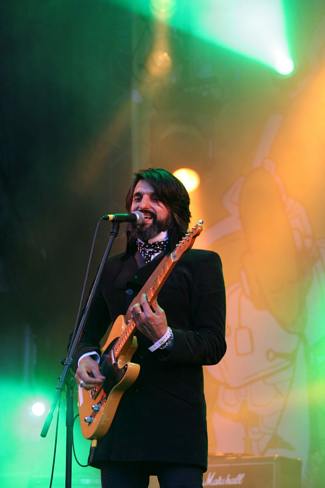 A musician with long hair and a beard performs on stage at the AB 2008 festival, playing an orange electric guitar. Wearing a dark suit and patterned scarf, he sings into a microphone, illuminated by green and yellow stage lights. In the background, there are speakers and colorful Positivus stage decorations.