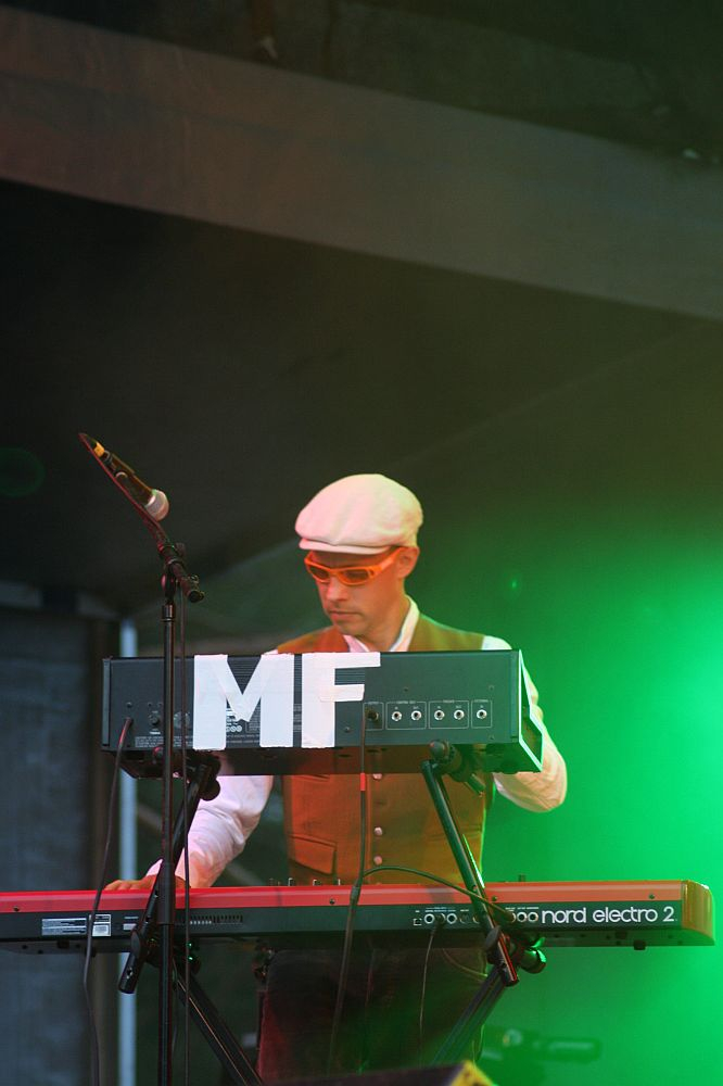 A musician wearing a white cap, sunglasses, and a brown vest stands behind a Nord Electro 2 keyboard on stage at Positivus. To the left is a microphone. The stage is illuminated with green lighting, and the letters "MF" are prominently displayed on the keyboard setup.