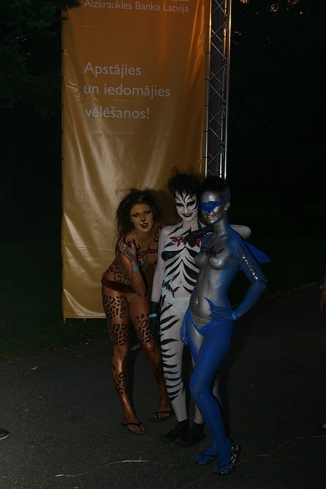 Three people in detailed body paint stand together outdoors at night. The person on the left is painted as a leopard, the middle as a tiger, and the right as a blue dolphin. Behind them is a large vertical yellow banner with Latvian text. The dark park setting suggests this could be Positivus AB 2008.