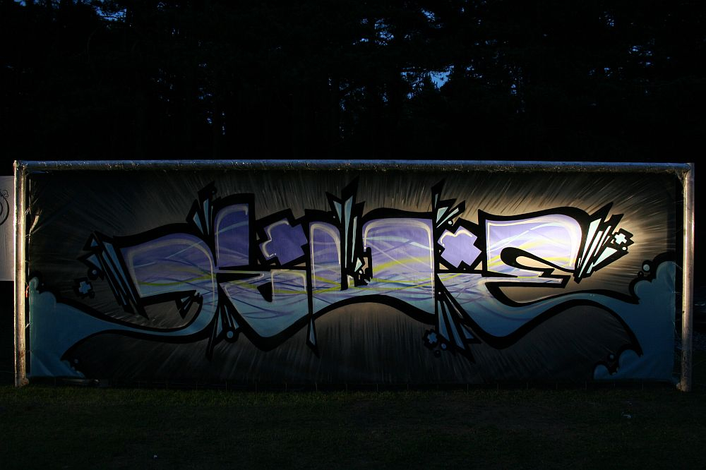 A metal fence displays vibrant graffiti with stylized letters and abstract designs in shades of blue, purple, and white. The graffiti, reminiscent of Positivus festival art, is illuminated against a dark, forested background. The colors and lines stand out prominently above the grassy ground beneath.