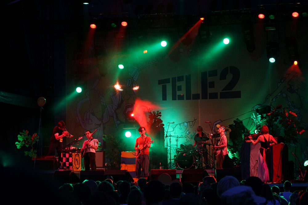 A band performing on a stage under colorful lights, featuring red and green hues, reminiscent of Positivus. Six musicians play various instruments, including guitars, a keyboard, and drums. A large backdrop with the "TELE2" logo and abstract designs is visible behind them. An audience is in the foreground.