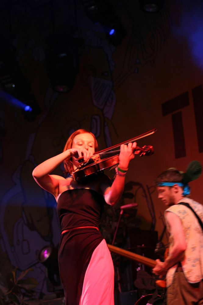 At Positivus, a woman passionately plays the violin on stage, dressed in a black and pink outfit. Behind her, a man with a blue headband plays a guitar. The background, featuring colorful artwork from AB 2008, is lit with vibrant stage lights, creating an energetic atmosphere.