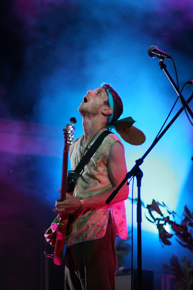 A person stands on stage under vibrant blue and purple lighting at Positivus 2008, playing an electric guitar. They are wearing a sleeveless shirt and a headband with a large feather attached. The person appears enthusiastic, looking upwards with an open mouth, and there are microphones and plants in the background.