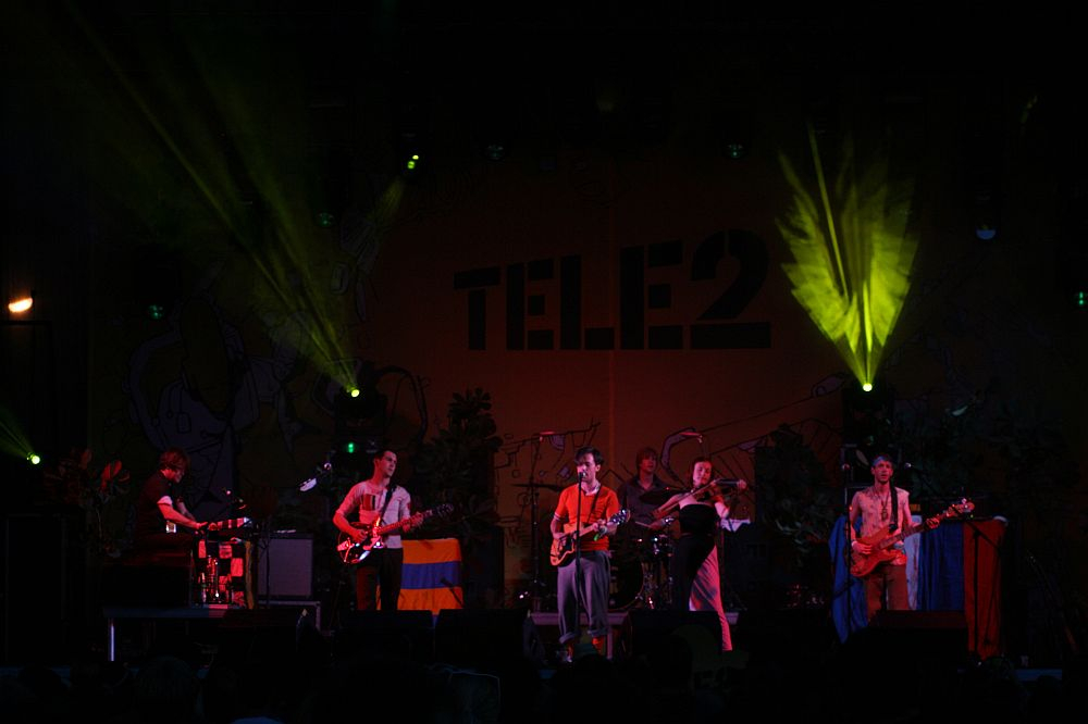 A band is performing on stage under green and yellow spotlights at Positivus 2008. The background features a "TELE2" banner. Musicians are playing keyboards, guitars, and drums with one draped in a colorful flag. The stage, adorned with plants, creates an atmospheric setting for the event.