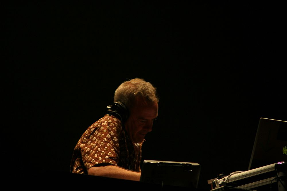 At Positivus 2008, a person with headphones stands at a DJ table in a dark setting. They are wearing a patterned shirt and appear focused on the equipment in front of them, which includes a laptop and other DJ gear. The background is dark, emphasizing the person and the equipment.