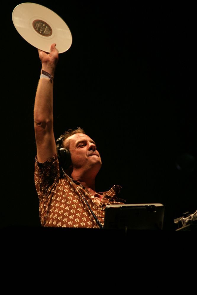 A DJ with headphones holds up a white vinyl record high in the air at Positivus. He wears a patterned shirt and an expression of triumph and concentration. The background is dark, highlighting the DJ and creating a dramatic atmosphere. There is DJ equipment in front of him, reminiscent of AB 2008 energy.
