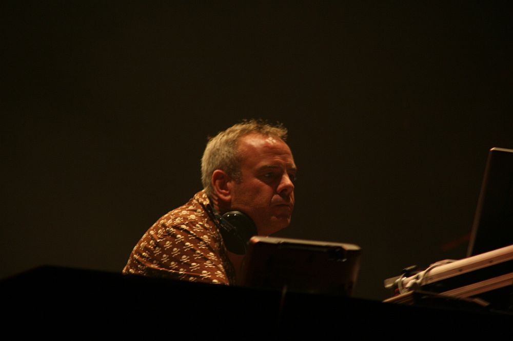 A person with short gray hair and wearing a patterned shirt is sitting in front of electronic equipment, possibly DJing at Positivus. They have headphones around their neck and appear focused on the task. The background is dark, creating a moody atmosphere with minimal lighting on the person's face.
