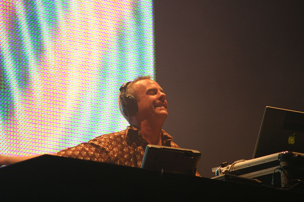 A person smiling broadly while performing as a DJ at Positivus. They are wearing headphones and standing behind a DJ console with a laptop featuring an Apple logo. The background is illuminated with vibrant, colorful lights. The person appears to be enjoying the moment during the performance.