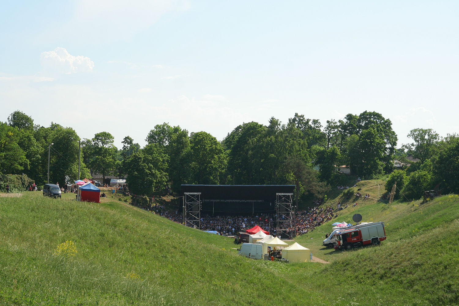 A large outdoor concert stage is set up in a grassy, wooded area. Crowds are gathered in front of the stage, ready for the Punk Laulupidu festival. Several tents and a red truck, possibly emergency services, are positioned to the right of the audience. The sky is clear and sunny, and the surrounding trees are lush and green.