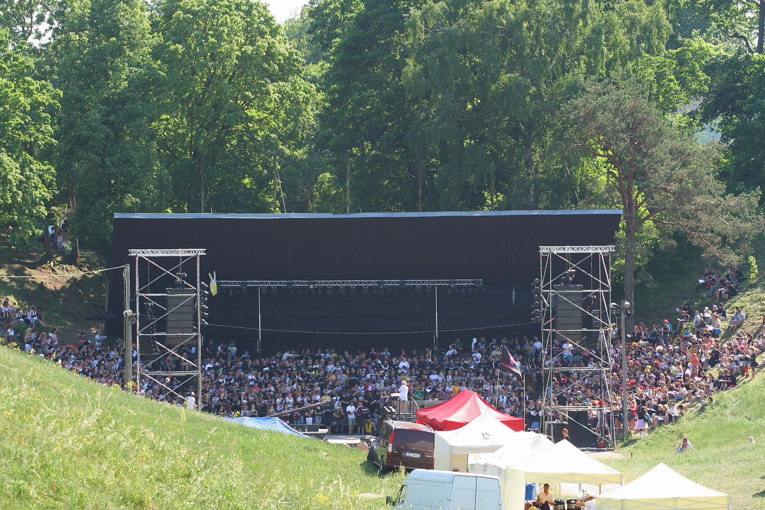 A large crowd gathers in a natural amphitheater with lush green trees in the background for Punk Laulupidu. The audience is seated in front of a stage with a large black backdrop, flanked by metal scaffolding towers. Several tents and vehicles are visible in the foreground, indicating an outdoor punk festival.
