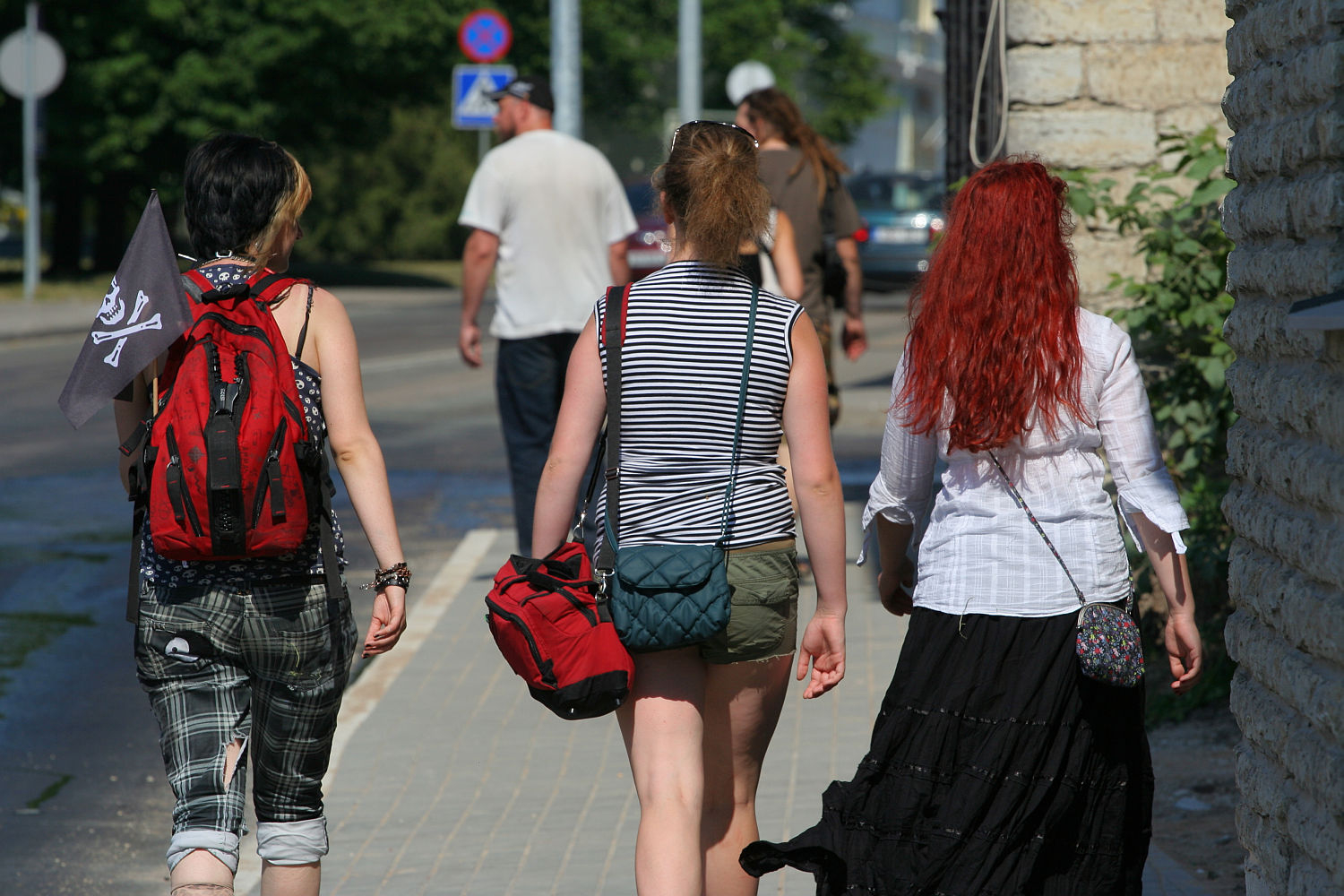 Three people walking down a sunny sidewalk. The person on the left, with short hair and a red backpack, carries a black flag that gives off a punk vibe. The middle person wears a striped tank top and khaki shorts. On the right, someone with long red hair sports a white blouse and black skirt. Trees and a beige building are in the background, reminiscent of Laulupidu gatherings
