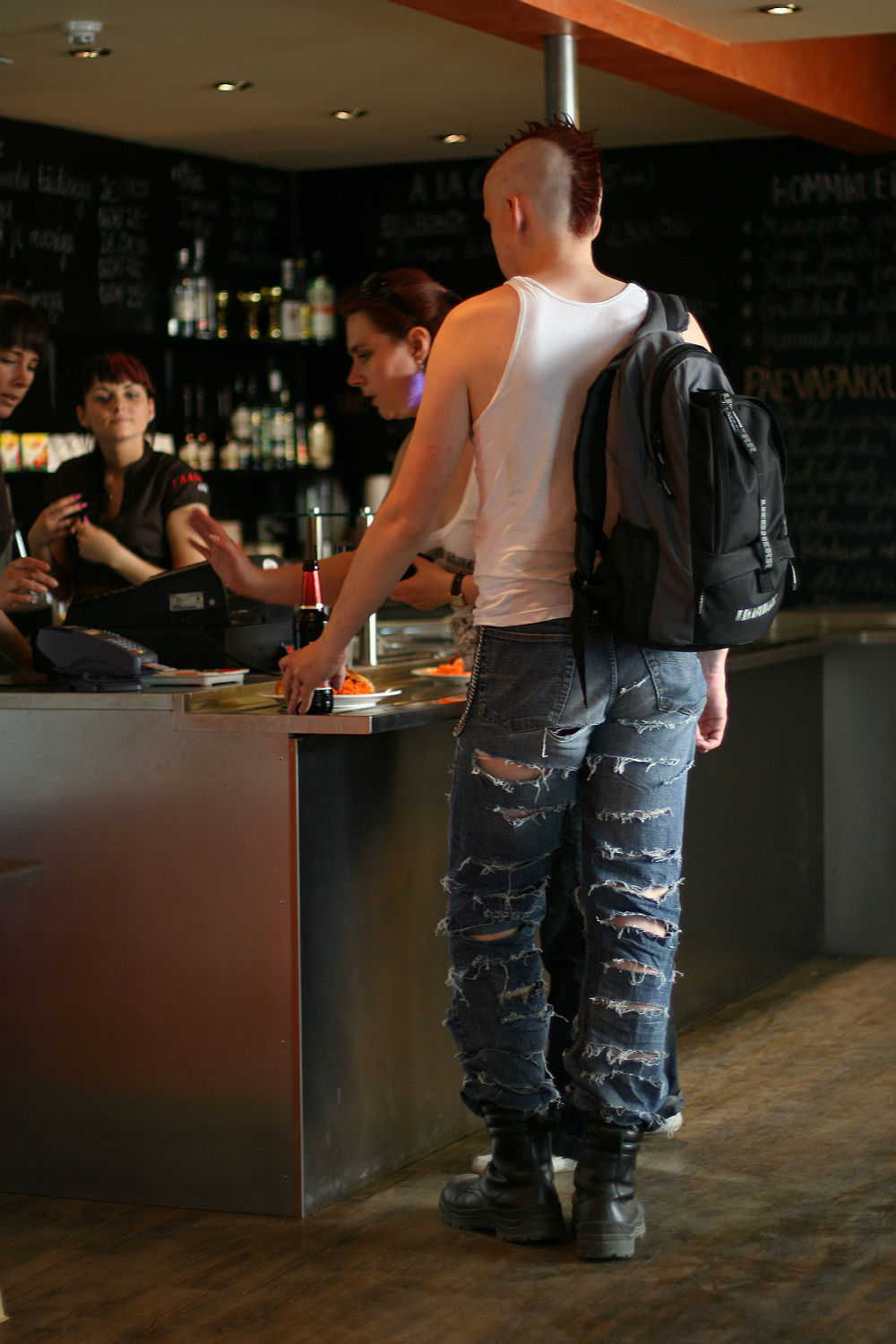 A punk with a mohawk haircut, wearing a white tank top, ripped jeans, black boots, and a black backpack, stands at a bar counter. They are holding a bottle and interacting with one of the three people behind the counter. The bar has shelves with bottles and a chalkboard with handwritten text.