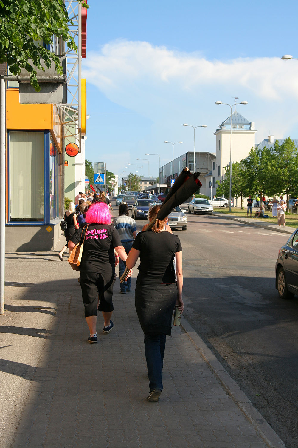A group of people walk on a sidewalk next to a street, some wearing black punk outfits. One person with a pink wig has text on their back. Another person carries a long, rolled-up item on their shoulder. Cars are parked along the road; buildings and trees line the street under a clear blue sky.