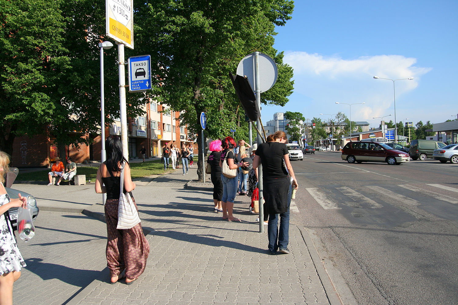 A group of people, some casually dressed, stand at a bus stop on a sunny day. Among them is a punk with vibrant hair, adding flair to the scene. Bright green trees provide shade as pedestrians walk along the sidewalk near a pedestrian crossing. Buildings and a bus stop sign with schedule information are also present.