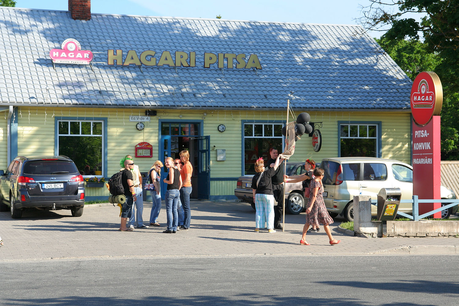 A group of people are gathered in front of a yellow and green building with a sign that reads "Hagari Pitsa." Parked cars are visible nearby. One person, looking punk, is holding black balloons. A person with flowers walks by on the sidewalk. A bright blue sky and greenery form the background.