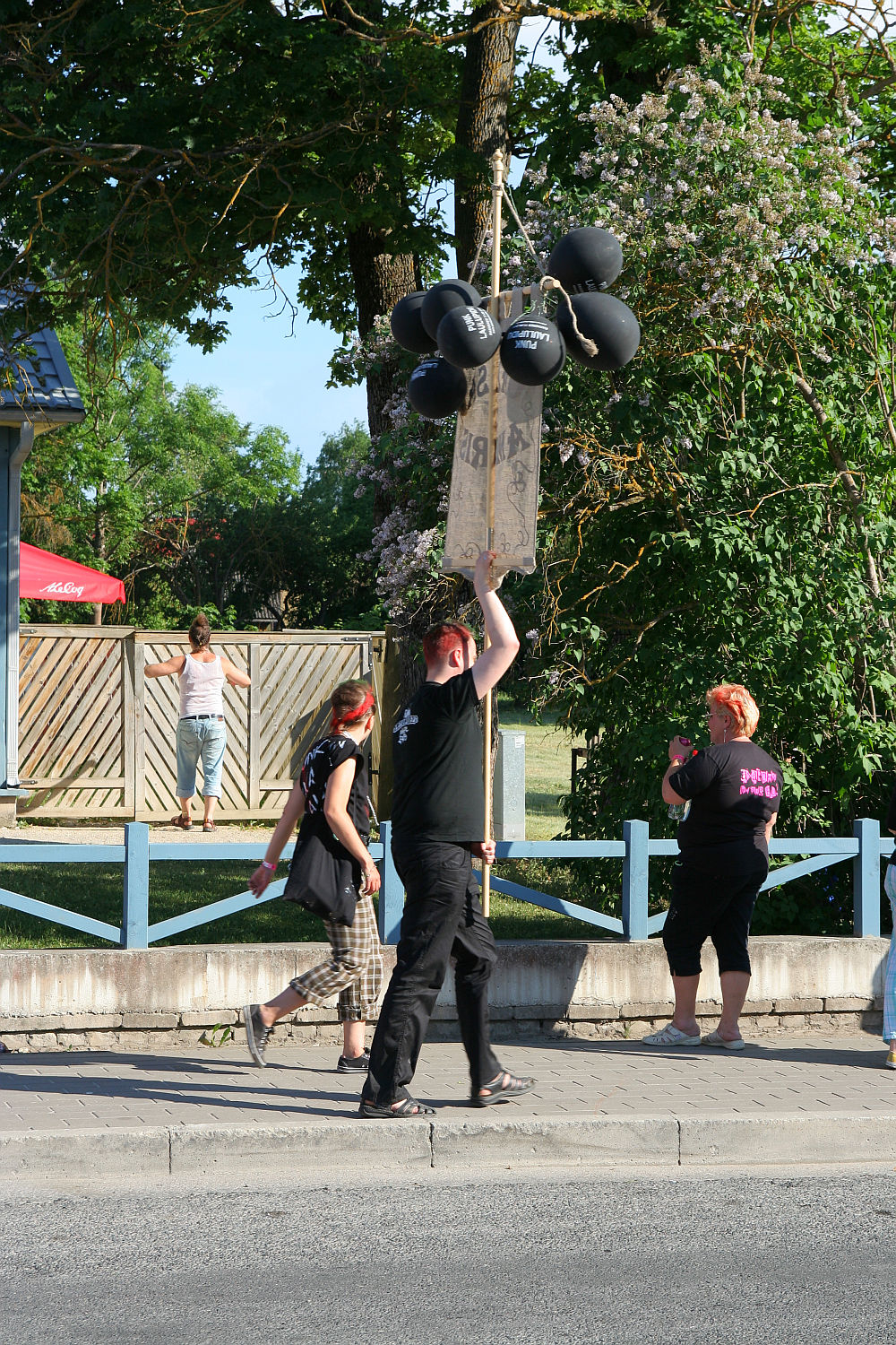A group of people dressed in black, embodying a punk vibe, stand on a sidewalk near wooden fencing. One person holds balloons and a sign. Another person, with orange hair, takes a photo. Trees and greenery fill the background, and a child sits on the fence. The scene takes place on a sunny Laulupidu day.