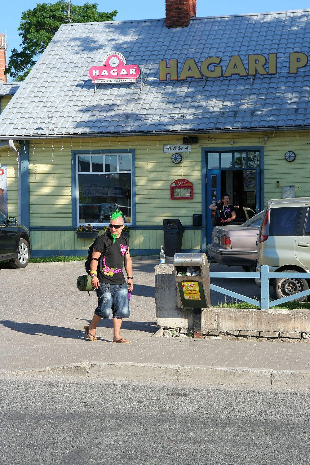 A punk sporting a black t-shirt, denim shorts, green mohawk, and neon pink sunglasses strolls down the sidewalk. He's carrying a cylindrical bag and a plastic bottle. Behind him, the two-story yellow building of "Hagari Pub" with its grey tiled roof stands out. A tan car is parked nearby.