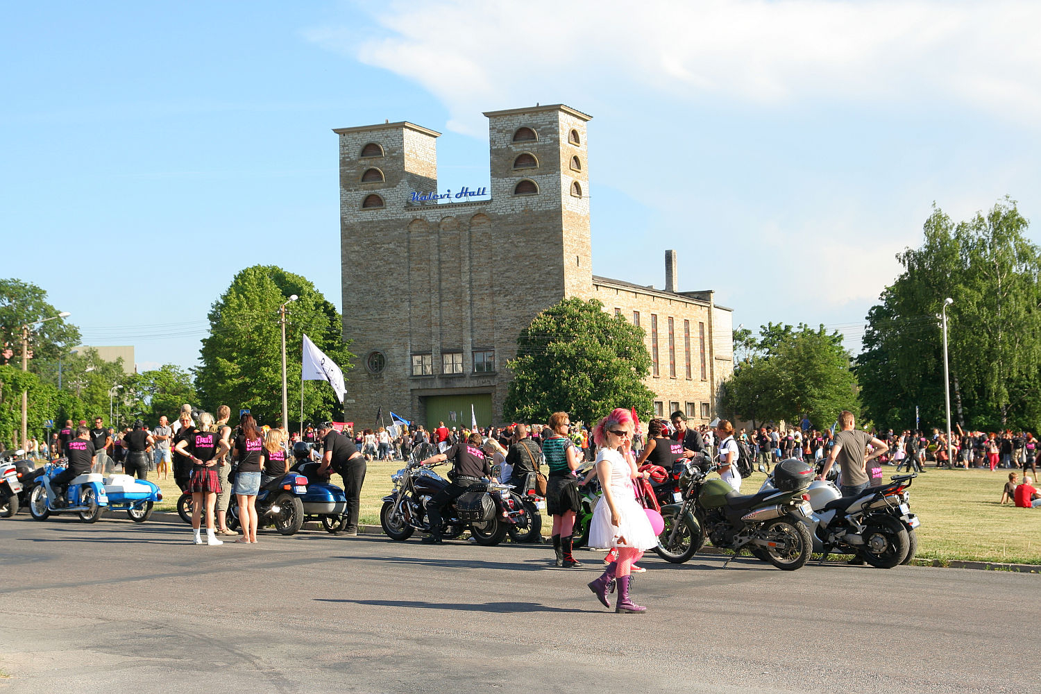 A large gathering of people is taking place outdoors near a historic building with twin towers. Multiple motorcycles are parked, and individuals are socializing. A child in a colorful outfit walks in the foreground, adding to the vibrant ambiance reminiscent of a festive Punk Laulupidu event under clear skies.