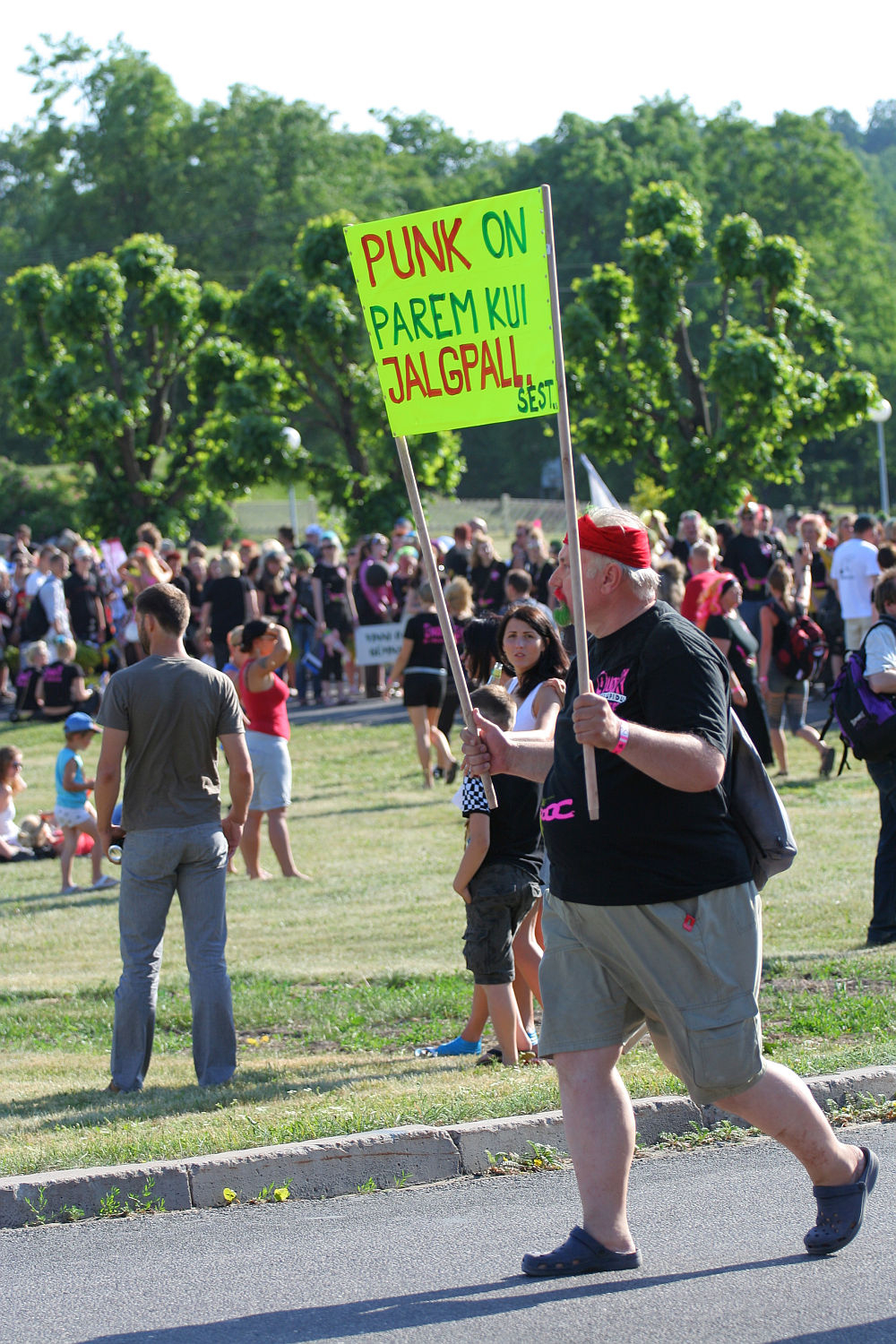 A person in a black shirt and red bandana is walking in a park holding a bright green sign with text in a foreign language. The person, seemingly part of the colorful Punk Laulupidu celebration, is among others gathered outdoors on a sunny day. Trees and grassy areas are visible in the background.