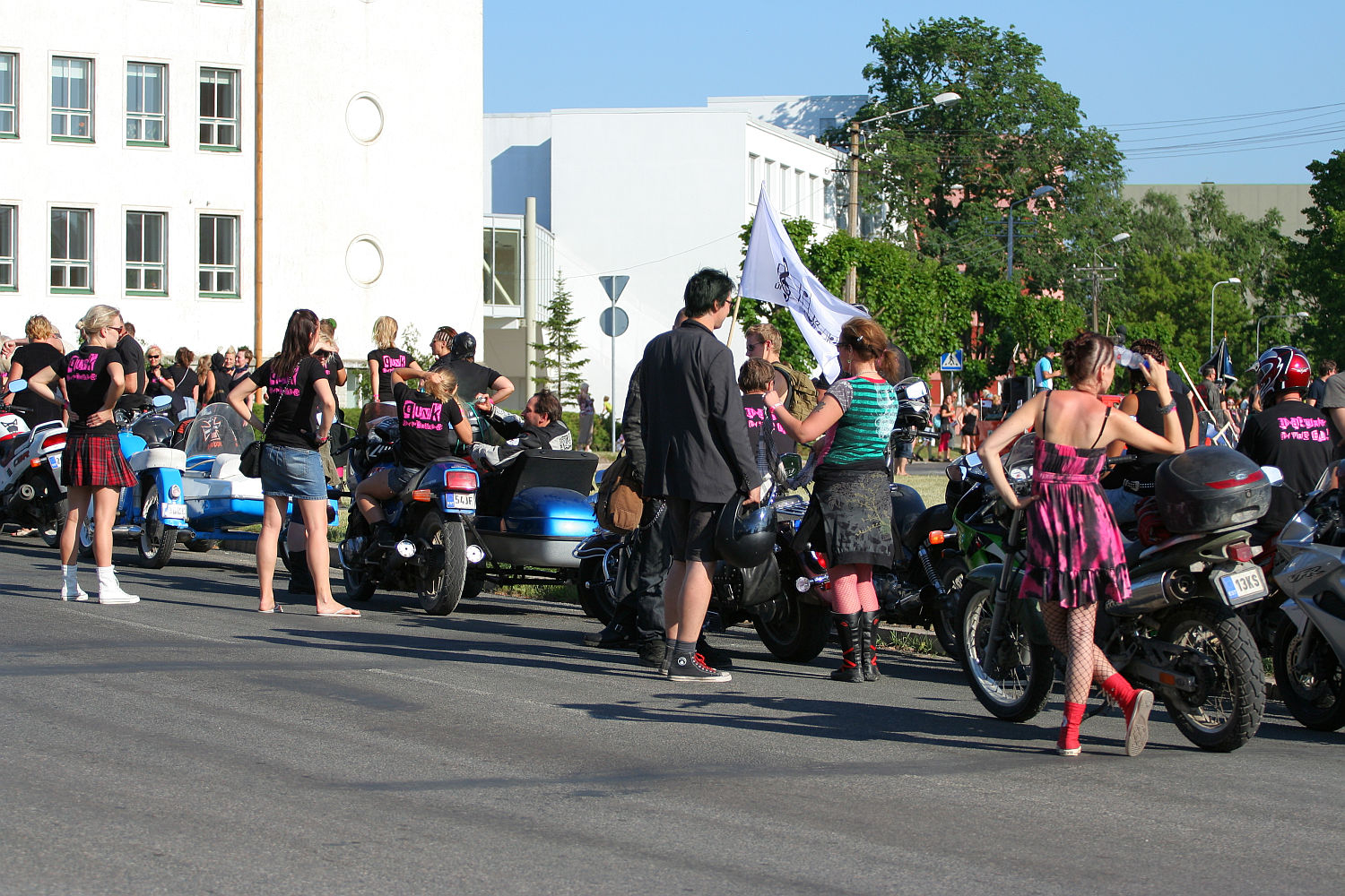 A group of people, mostly wearing black and pink punk-style outfits, gather around motorcycles on a sunny day. Some are sitting on the bikes while others stand nearby. Buildings and trees are in the background, and a few people hold a white flag. The scene has a vibrant, festive Laulupidu atmosphere.