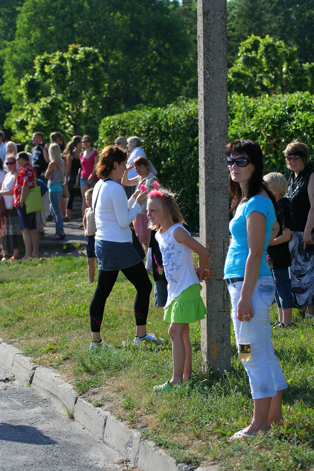 A group of people standing outdoors on a sunny day, gathered along a curb and green hedge, perhaps enjoying the vibrant atmosphere of Laulupidu. In the foreground, a girl in a white top and green skirt stands next to a woman wearing sunglasses and holding a beverage. Other adults and children are spread out along the path.
