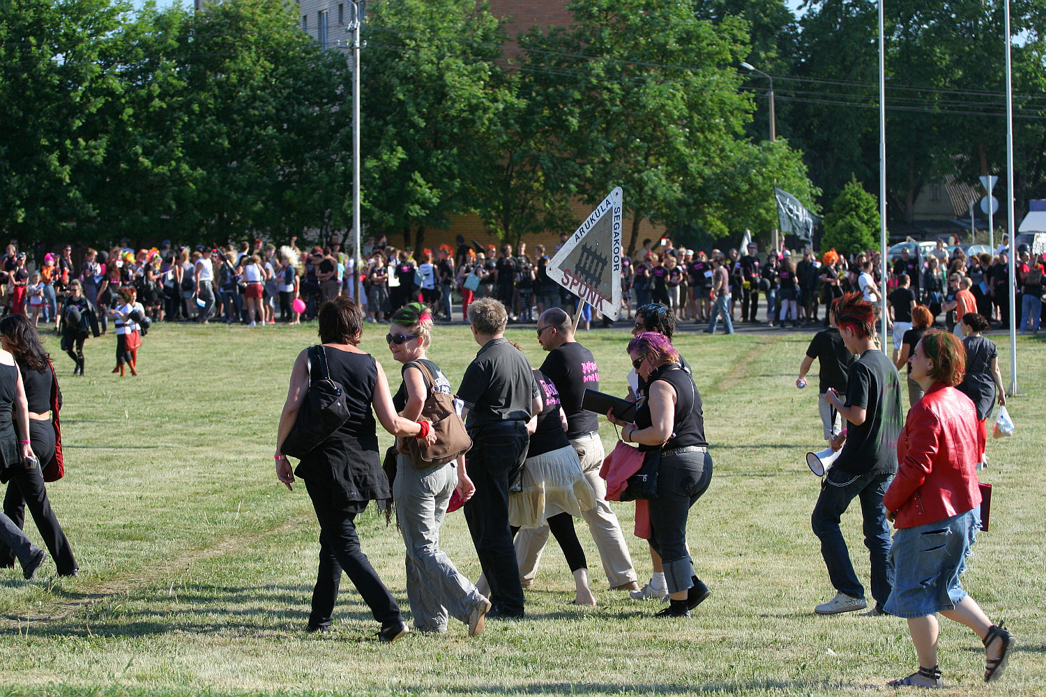 A large outdoor gathering with numerous people walking and standing on a grassy field. Most individuals are dressed in black clothing, reminiscent of a punk festival. Verdant trees and a brick building are visible in the background. A large banner and several flags, like those seen at Laulupidu, are held by participants in the distance.